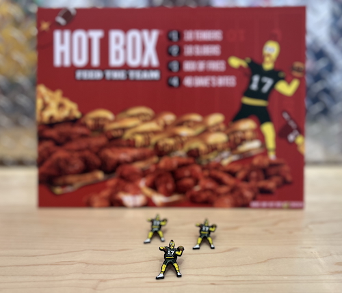 Limited-edition enamel pins of the Dave's Hot Chicken mascot wearing a green and gold football uniform, throwing a chicken slider as if it were a football. The pins are posed in front of a Hot Box menu at a Dave's Hot Chicken restaurant.