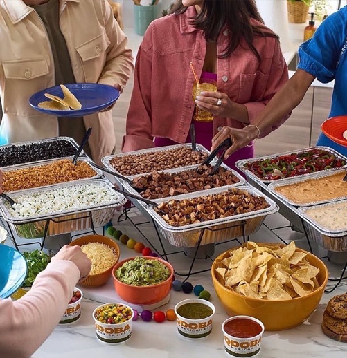 Guests serving themselves at a colorful Mexican buffet catered by QDOBA Catering, featuring build-your-own tacos with various meats, toppings, salsas, queso, guacamole, and tortilla chips.